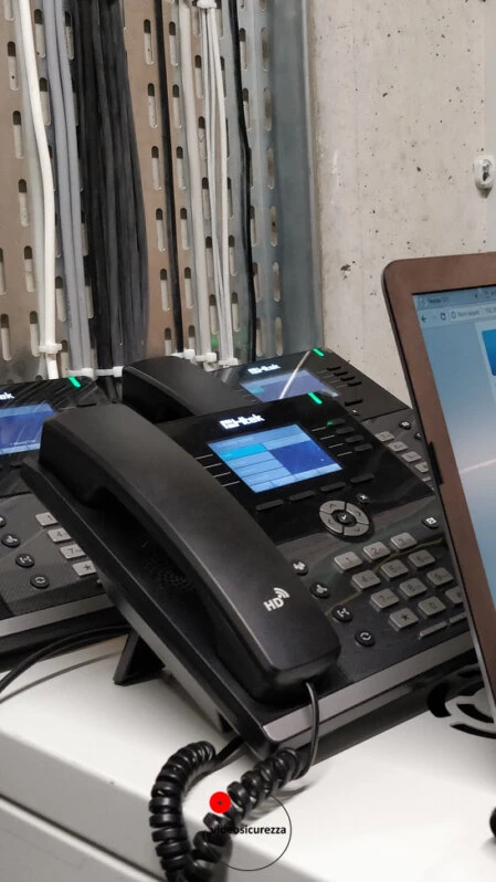 Telefoni VoIP da scrivania in configurazione su banco tecnico con notebook, cablaggio visibile sullo sfondo