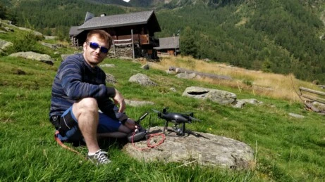Pilota di drone seduto su un prato alpino pronto al decollo, con controller e drone posati su roccia, sullo sfondo una baita in legno tra i monti